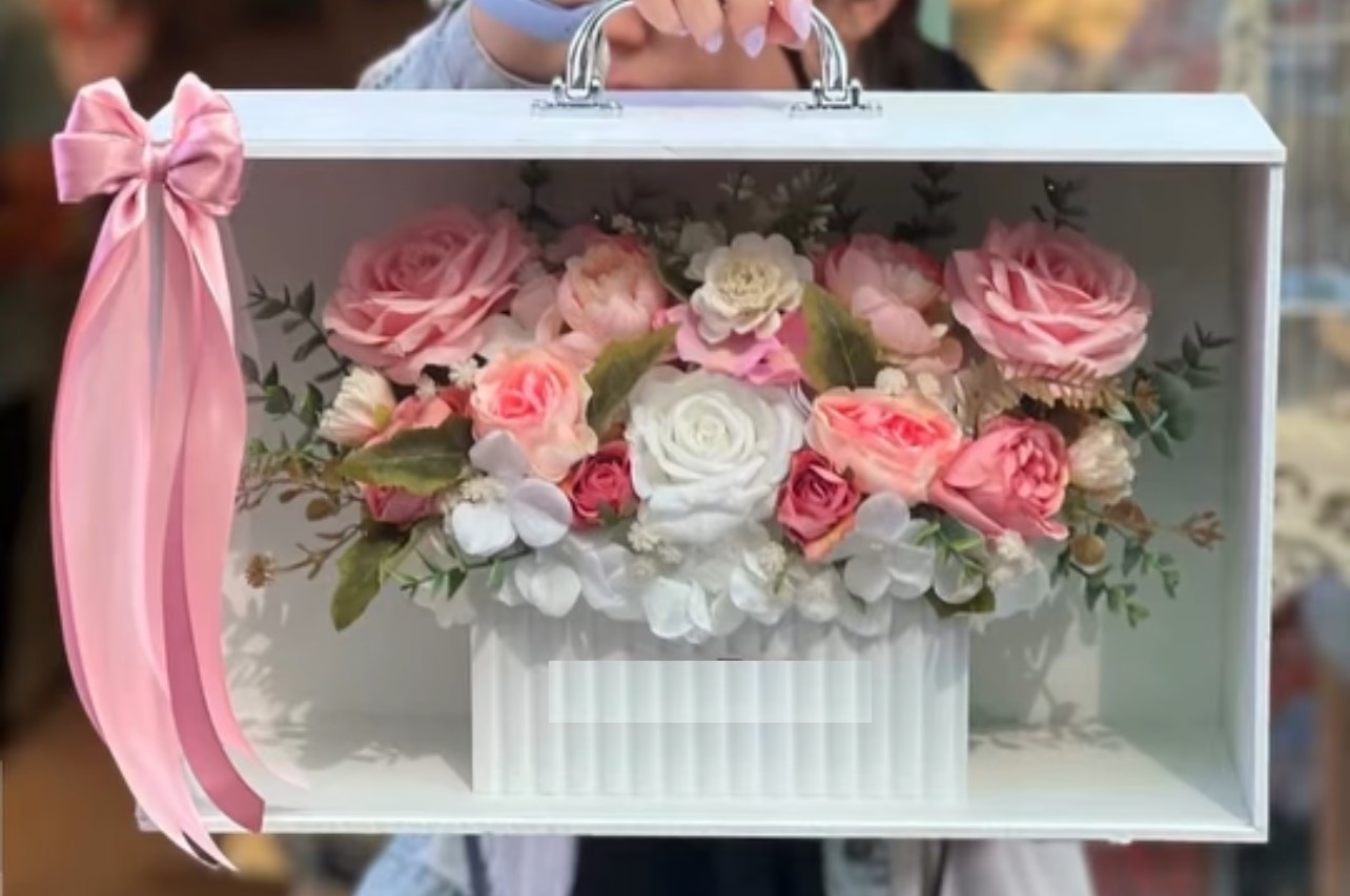 باکس چمدانی سفید با نمای شیشه‌ای، پر شده از گل‌های رز مصنوعی به رنگ‌های صورتی روشن، سفید و گلبهی با روبان صورتی ساتن - برند سام فلاور.
